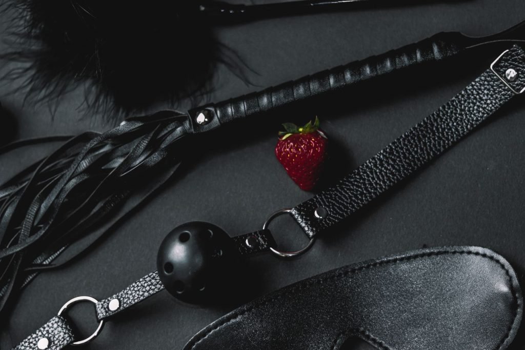 A set of black leather BDSM accessories featuring a ball gag and whip, contrasted with a fresh strawberry.